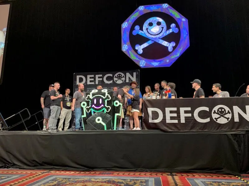A group on the DEF CON stage beside a glowing skull-and-crossbones display