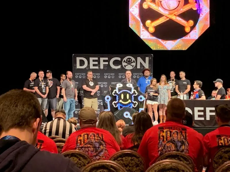 The same DEF CON stage seen from the audience, with goons in red shirts in the front rows