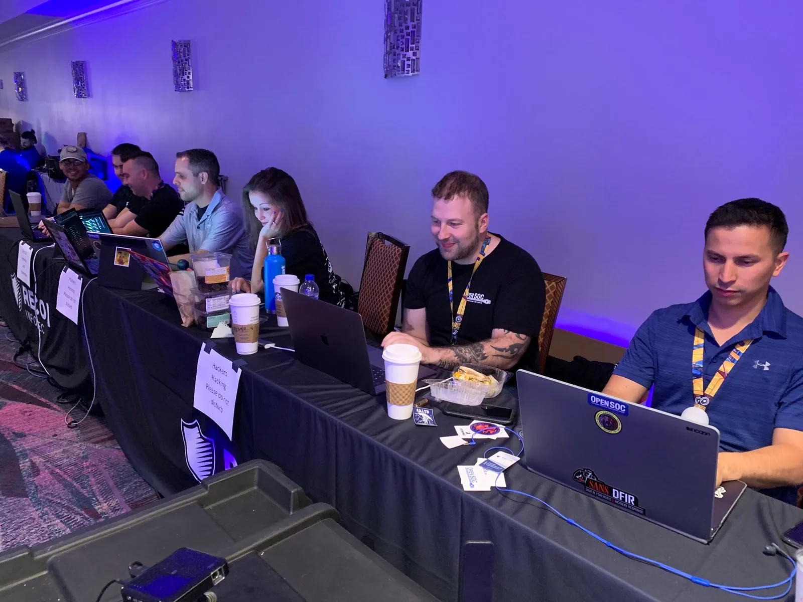 Team members at laptops along a Recon InfoSec table with Hackers Hacking Please do not disturb signs