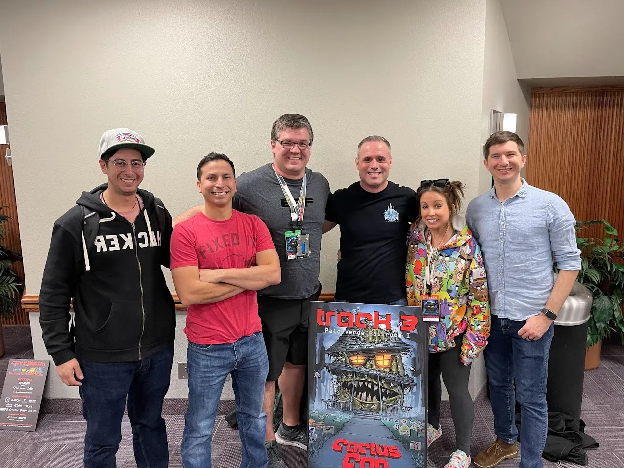 Six people smiling beside the CactusCon Track 3, Palo Verde Ballroom I sign